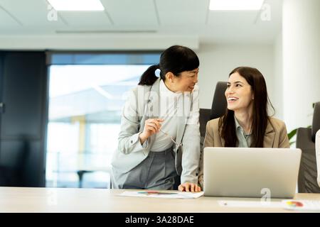 In a contemporary office setting, two women engage in a cheerful conversation, exchanging ideas and smiles while working on a laptop, creating a posit Stock Photo