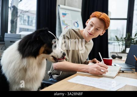 A young man with red hair writes notes in an office, his dog sitting beside him. Stock Photo