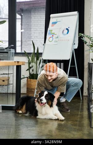 A young red haired man bends down to pet a happy dog while working in a stylish office environment. Stock Photo