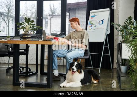 Focused young man with red hair engages in work at a stylish office while his dog relaxes nearby Stock Photo