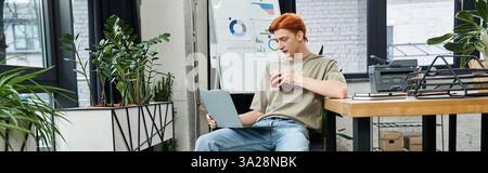 A dedicated young man with red hair focuses on tasks while enjoying a drink in a stylish workspace. Stock Photo