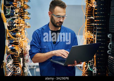 Holding laptop. Young man is working with internet equipment and wires in server room. Stock Photo