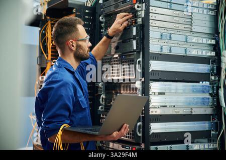 Using the laptop. Young man is working with internet equipment and wires in server room. Stock Photo