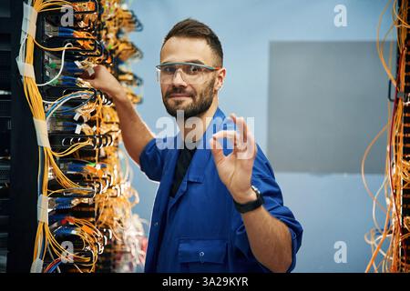 Okay or fine hand gesture. Young man is working with internet equipment and wires in server room. Stock Photo