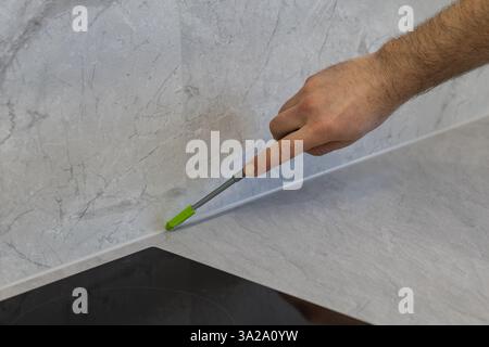 Hand of worker using silicone sealant applicator to fill gap between kitchen countertop and wall with marble effect. Home renovation, sealing process Stock Photo