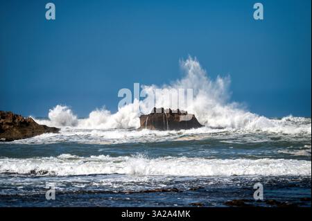 Waves crash against rocky outcrop along the coast of Essaouira, Morocco under bright blue sky Stock Photo
