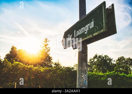 A weathered street sign reading 'Zur Loamgrui' stands at the edge of a vineyard, with the sun setting in the background, casting a warm glow over the Stock Photo