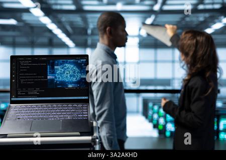 Laptop in front of data center engineers in blurry background deploying artificial intelligence tools. Close up of AI coding on notebook screen next to technicians inspecting server clusters Stock Photo