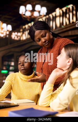 Two preteens girls and their teacher collaborating on schoolwork at the library, focused on academic success and learning development. Students collect extra information with private lessons. Stock Photo