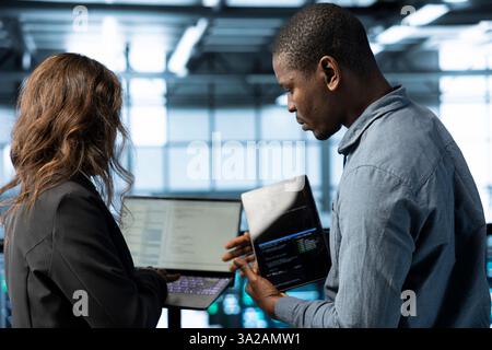 System administrators in data center using laptop and tablet, integrating green technologies. IT specialists in server room using devices to implement sustainable energy solutions Stock Photo