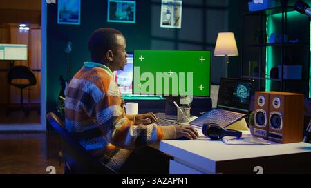 Black person programming neural network code with a green screen on computer to build databases. System engineer working from home while looking at data center security servers. Camera B. Stock Photo