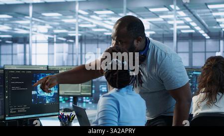 Manager coming to technician desk to give instructions on doing maintenance on artificial intelligence neural networks. Supervisor overseeing worker using AI system software, camera A Stock Photo