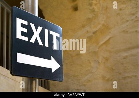Exit sign with arrow pointing right, white letters on black background with stone wall in the background, Valetta, Malta. Stock Photo