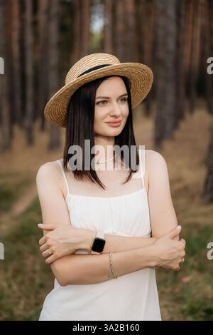 A young woman poses elegantly in a flowing white dress and a stylish straw hat surrounded by tall, lush trees Stock Photo