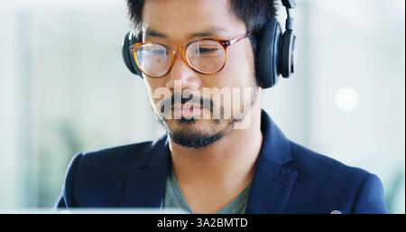 Developer, headphones or man with glasses in office for technology, podcast playlist or coding. Face, music and Asian programmer on computer with Stock Photo