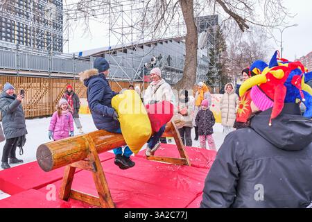 Yekaterinburg, Russia - February 26, 2025: People participate in traditional pillow fight game on wooden beam during Maslenitsa festival. Spectators, Stock Photo