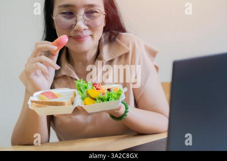 Cropped view of a black haired adult female siting at wooden table eating apple with salad and looking her laptop. Selective focus. Stock Photo