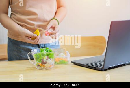 Cropped of woman preparing healthy food in kitchen while adding pumpkin and apple to salad and watching tutorial on laptop. Selective focus. Stock Photo