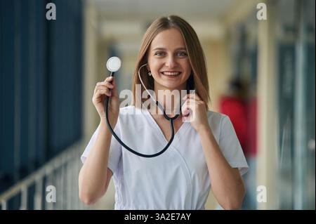 Using stethoscope. Female doctor in white coat is in the hall. Stock Photo