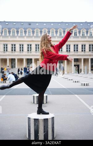 A stylish woman in a red coat and black dress strikes an elegant ballet pose on a striped column at Palais-Royal, Paris. The historic architecture and surrounding people enhance the artistic. Stock Photo
