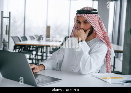 Working by using laptop. Successful Muslim businessman in traditional outfit in his office. Stock Photo