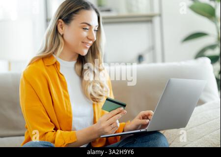 Happy caucasian woman in a yellow shirt sits on a cozy couch, smiling while using her laptop for online shopping, holds a credit card in her hand, entering banking information, paying bills, smiles Stock Photo