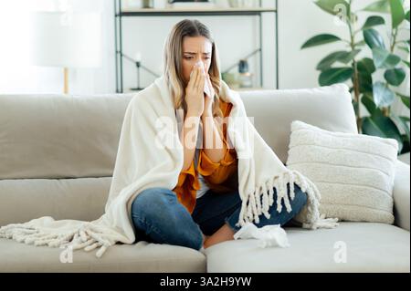 Upset unhealthy caucasian woman in a yellow shirt sits wrapped in a soft white blanket on a couch in living room, blowing her nose into a tissue, she looks unwell, showing symptoms of a cold or flu Stock Photo