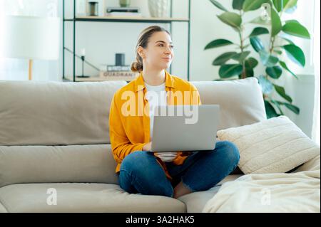 Relaxed caucasian woman in a yellow shirt and jeans sits comfortably on a sofa with a laptop, she looks into the distance with a thoughtful expression, enjoying home atmosphere in a cozy home, smile Stock Photo