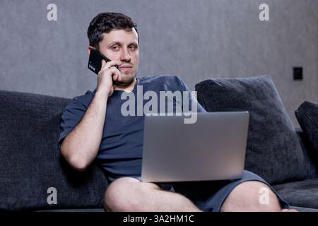 A man on a couch talks on a phone and uses a laptop Stock Photo