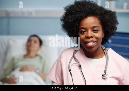 Portrait of female African American doctor smiling confidently with stethoscope, patient lying in bed in background, hospital setting showing care and professionalism Stock Photo