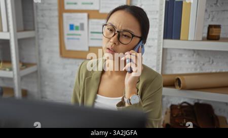 Woman talking on phone in modern office, looking focused, surrounded by graphs and shelves, wearing glasses and blazer, suggesting professional settin Stock Photo