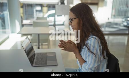 Young woman with brunette hair and glasses working on a laptop in a bright, modern office setting, smiling and engaging in an online meeting or video Stock Photo