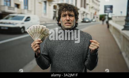 Hispanic man outdoors on city street displaying russian roubles with a frustrated expression, highlighting financial struggles or currency issues. Stock Photo