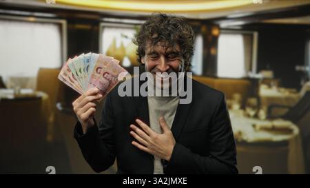 Man smiling indoors holding thai baht currency in a cozy restaurant setting, suggesting happiness and financial success. Stock Photo