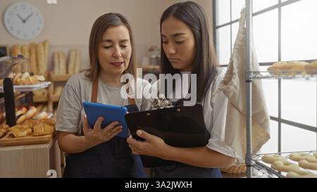 Women employees working together in a bakery analyzing data on tablet and clipboard indoors by shelves of bread Stock Photo