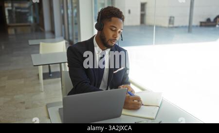 Young, handsome, african american man wearing a headset works attentively in a modern office, writing notes with a laptop and notebook on a desk. Stock Photo