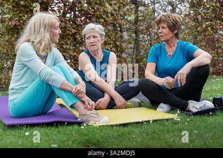 Three senior women sitting on yoga mats outdoors, engaged in a serious conversation. Concept of support, friendship, mental health, and community for Stock Photo