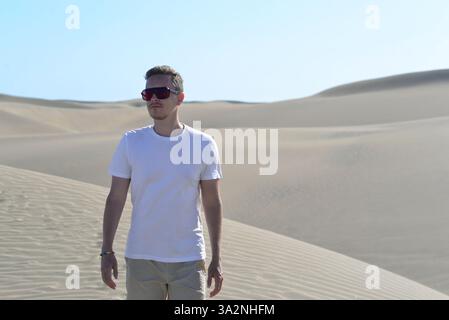 A stylish model portrait of a young man walking along a sand dune through barkhan in Maspalomas, Gran Canaria on a hot sunny day. Desert travel touris Stock Photo