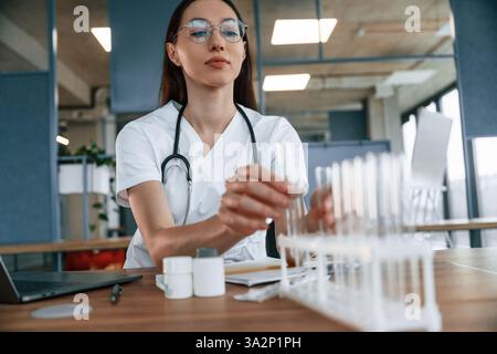 Woman scientist with stethoscope is sitting in lab and working with test tubes. Stock Photo