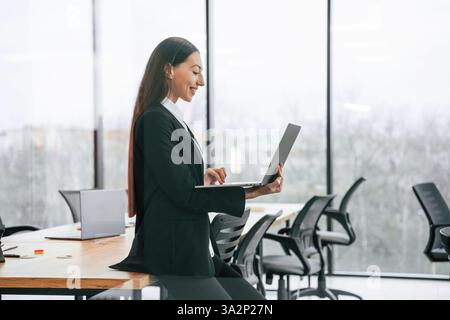 Side view. Standing and holding laptop. Businesswoman is indoors in the office. Stock Photo