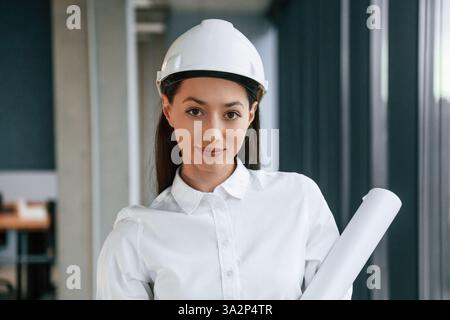 Portrait of female architect that is in hard hat and in white clothes indoors. Stock Photo
