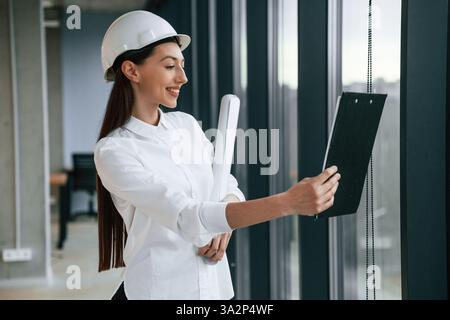 Plan and notepad in hands. Female architect in hard hat and in white clothes is indoors. Stock Photo