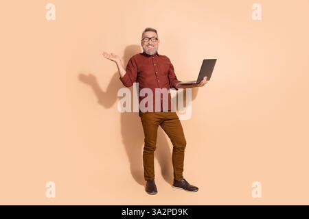 Smiling mature man with laptop gesturing confidently against beige background, showcasing gentlemanly charm Stock Photo