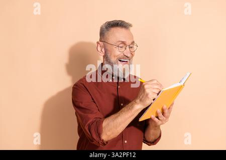 Handsome senior man writing notes in a notebook, appearing happy and content, on a beige background Stock Photo