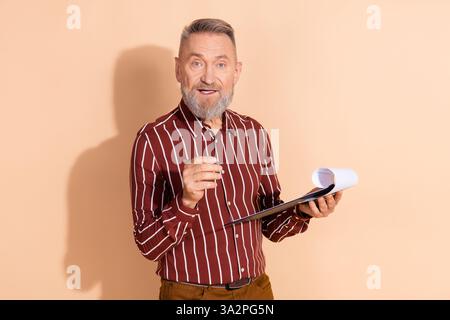 Smiling mature man using clipboard on beige background with striped shirt demonstrating cheerful professional demeanor Stock Photo