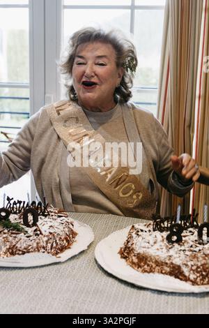 Happy senior woman singing and celebrating her 80th birthday, surrounded by two cakes with festive decorations. Stock Photo