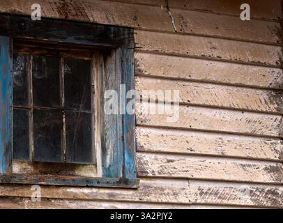 A weathered window with rustic blue frame on an aged wooden house, showcasing faded paint, peeling textures, and vintage charm. Stock Photo