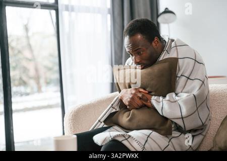 Sitting on sofa, holding pillow. Sick black man is at home. Stock Photo