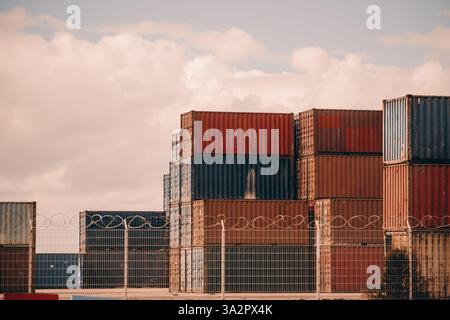 Stacked shipping containers in red and blue colors behind a barbed wire fence at an industrial storage yard. The containers show signs of rust and wea Stock Photo