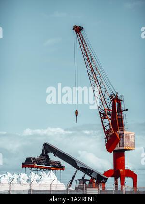 Industrial harbor crane lifting bulk cargo bags behind security fence, bright red crane structure, mechanical equipment, blue cloudy sky background. V Stock Photo